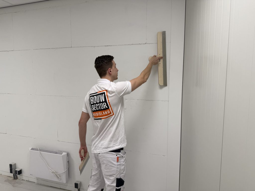 Stukadoor van Bouwsector Nederland zet een lange spaan hoog tegen een wand, met een kachel op de vloer